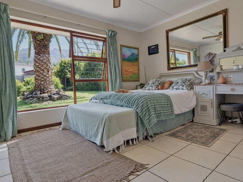 A bright bedroom featuring large windows with garden views, tiled flooring, a ceiling fan, and a built-in vanity unit.