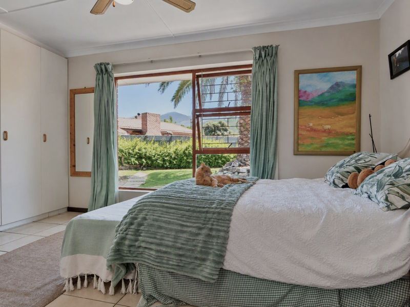 A bright bedroom featuring built-in cupboards, tiled flooring, a ceiling fan, and a large window with views of the garden.