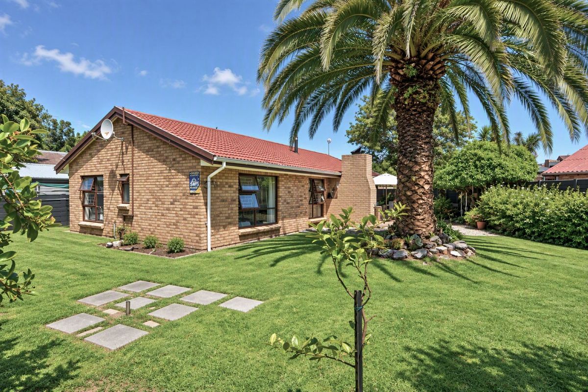 A well-maintained brick house exterior featuring a tiled roof, chimney, and established garden with a large palm tree.
