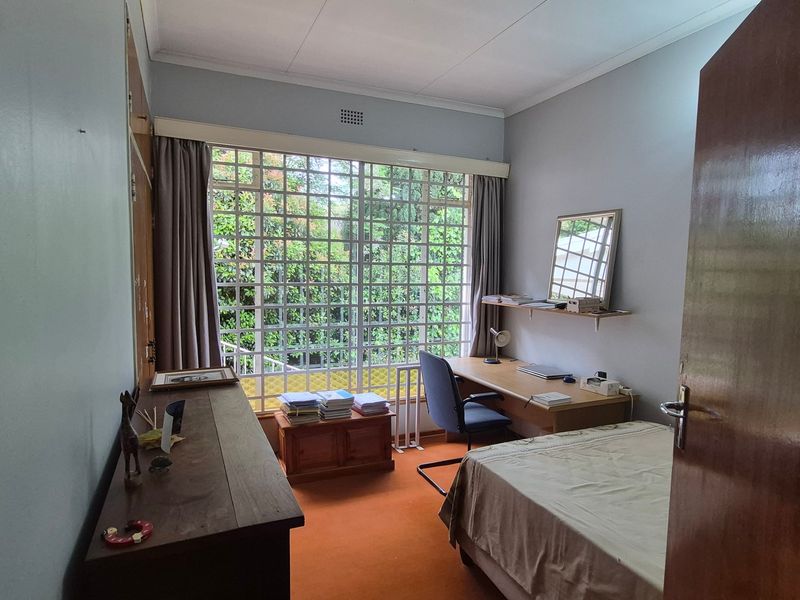 A functional bedroom featuring built-in cupboards, carpeted flooring, and a large window with a grid design, offering natural light and garden views.