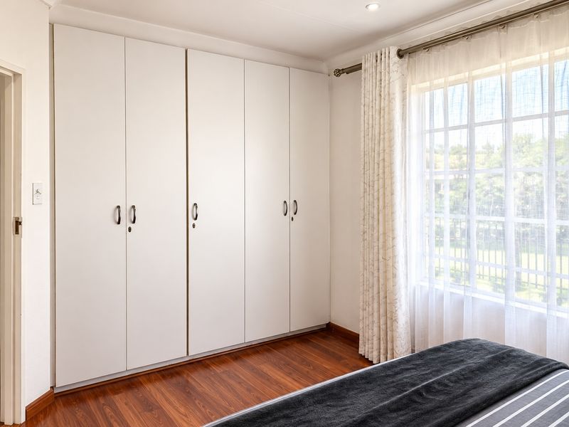 A well-maintained bedroom featuring floor-to-ceiling built-in cupboards, warm laminate flooring, and a window with security bars.