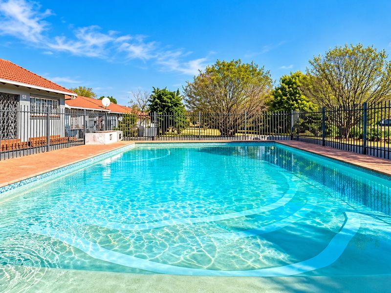 A well-maintained swimming pool area featuring clear water, brick paving, and secure metal perimeter fencing, set against a clear blue sky.