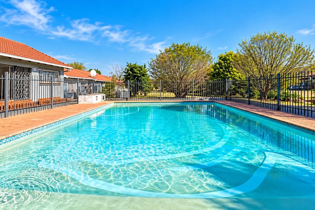 A well-maintained swimming pool area featuring clear water, brick paving, and secure metal perimeter fencing, set against a clear blue sky.
