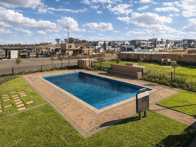 A well-maintained communal swimming pool area featuring brick paving, landscaped lawns, and a built-in brick bench within a residential estate.