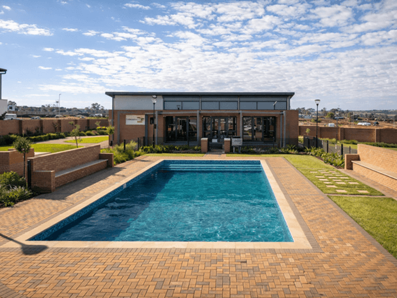 A well-maintained outdoor pool area featuring a rectangular swimming pool, brick paving, built-in seating, and landscaped lawn, adjacent to a modern clubhouse facility.