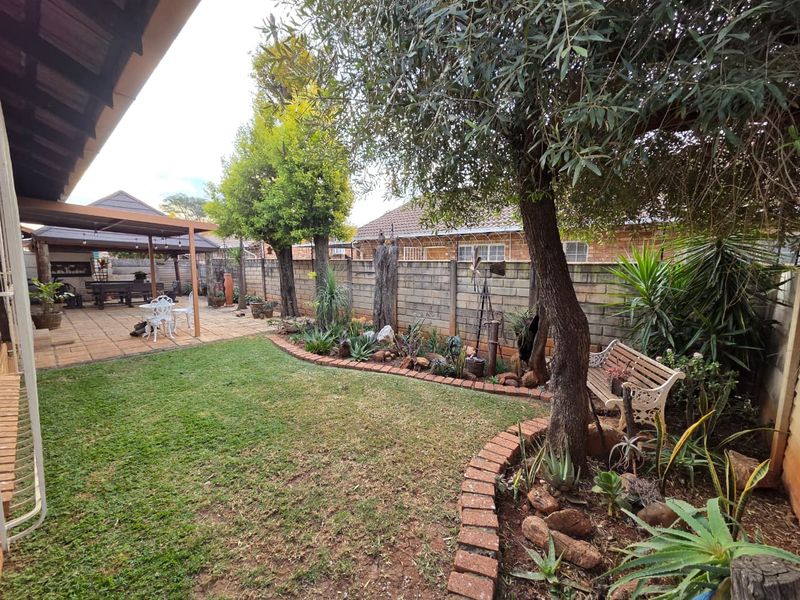 A well-maintained rear garden featuring a covered patio area, established trees, landscaped garden beds with brick edging, and secure perimeter walling.