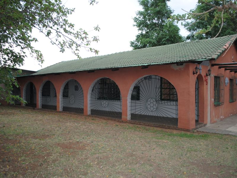 Rear view of a single-story house featuring an arched veranda, security gates, and a tiled roof, set within a garden area.