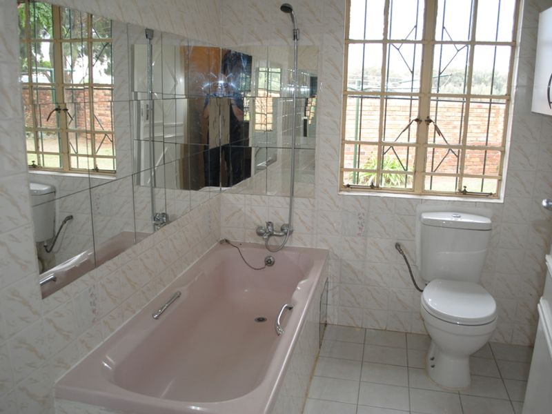 A dated bathroom featuring a pink built-in bathtub, white toilet, tiled finishes, and security bars on the window.