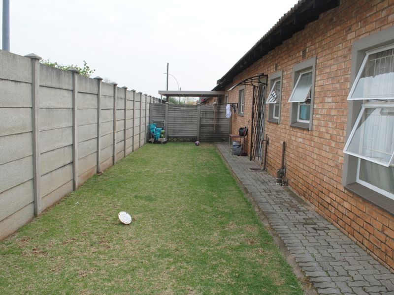 A rear exterior view of the townhouse featuring a brick facade, paved walkway, and a private lawn enclosed by a precast concrete boundary wall.