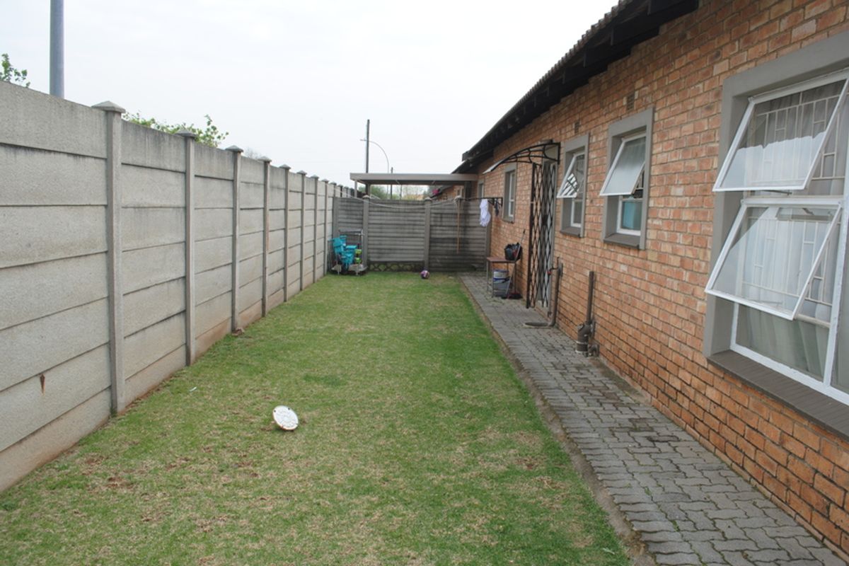 A rear exterior view of the townhouse featuring a brick facade, paved walkway, and a private lawn enclosed by a precast concrete boundary wall.