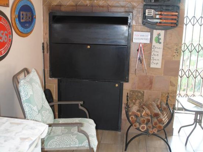 A covered patio area featuring a built-in braai, stone tile wall cladding, and ceramic floor tiles, secured by a metal gate.