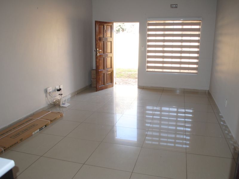 An empty, well-lit living room featuring neutral tiled flooring, a timber entrance door, and modern roller blinds.