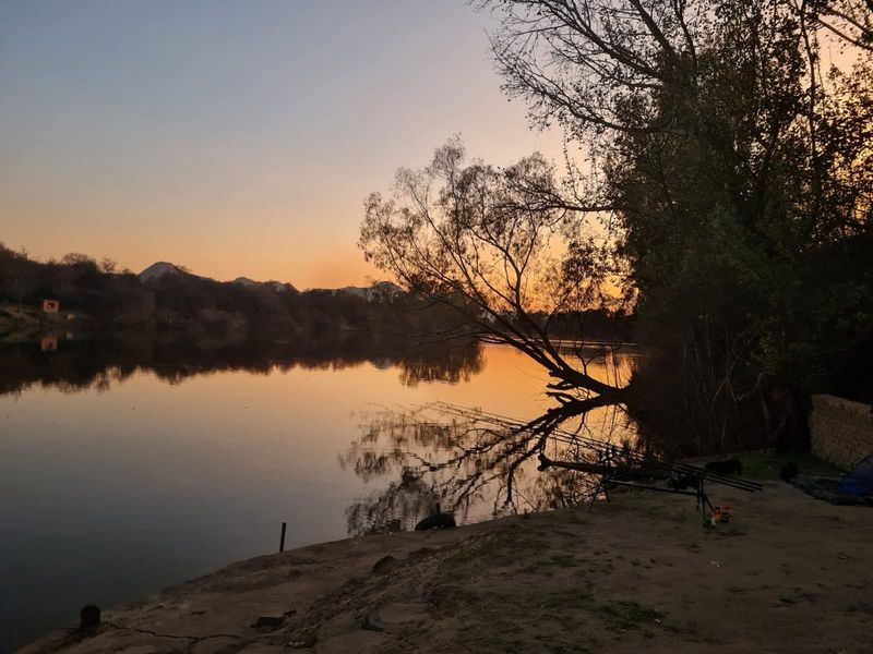 A scenic sunset view overlooking a calm river with natural landscape and mountain silhouettes in the background.