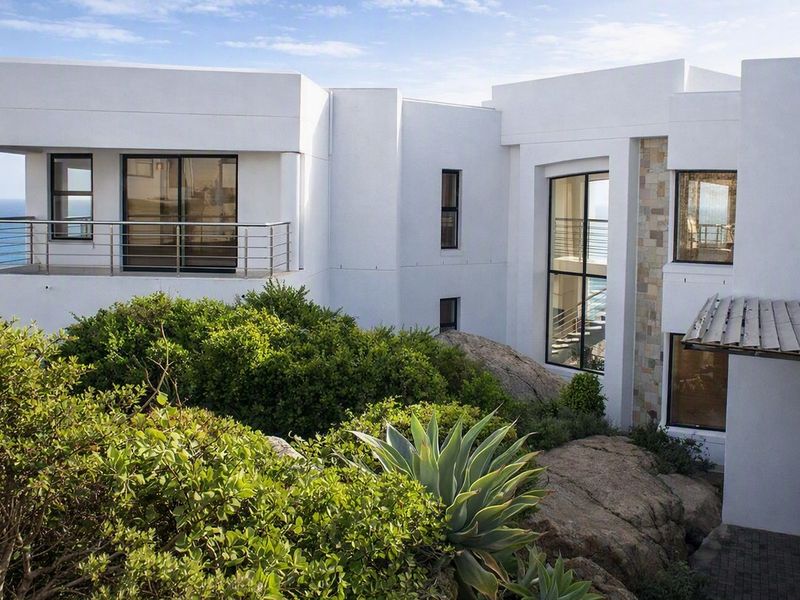 Modern multi-level residence featuring clean white facades, stone accents, expansive glass windows, and a balcony overlooking the coastal landscape.