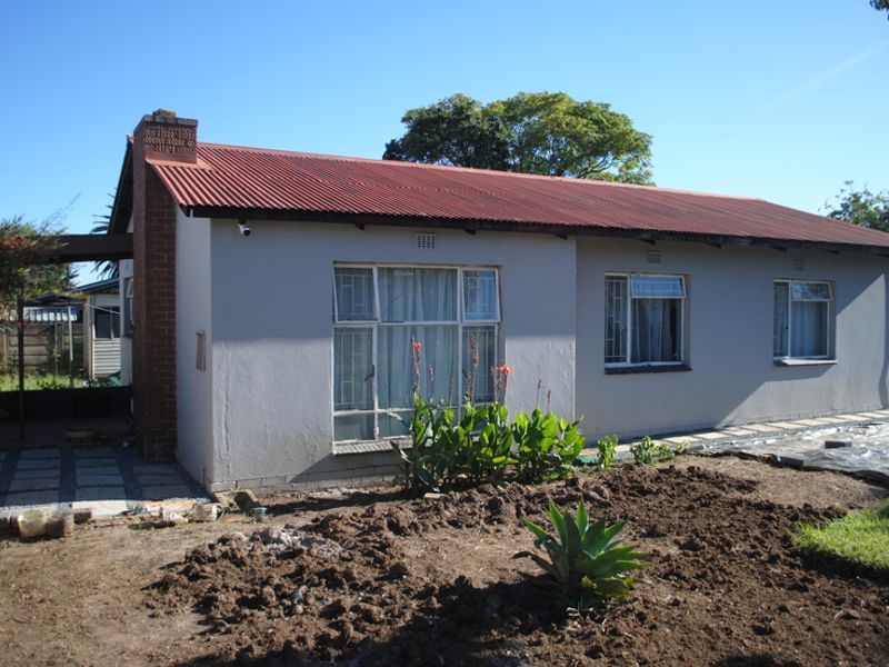 Rear view of a single-story house featuring a corrugated metal roof, brick chimney, and security bars on the windows, with an unlandscaped garden area.