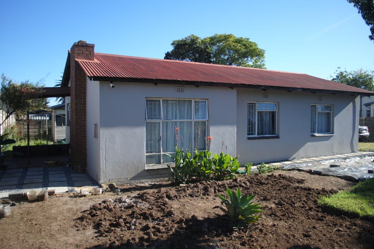 Rear view of a single-story house featuring a corrugated metal roof, brick chimney, and security bars on the windows, with an unlandscaped garden area.
