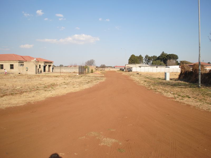 A view of an unpaved residential street showing surrounding properties and perimeter fencing in a developing neighborhood.