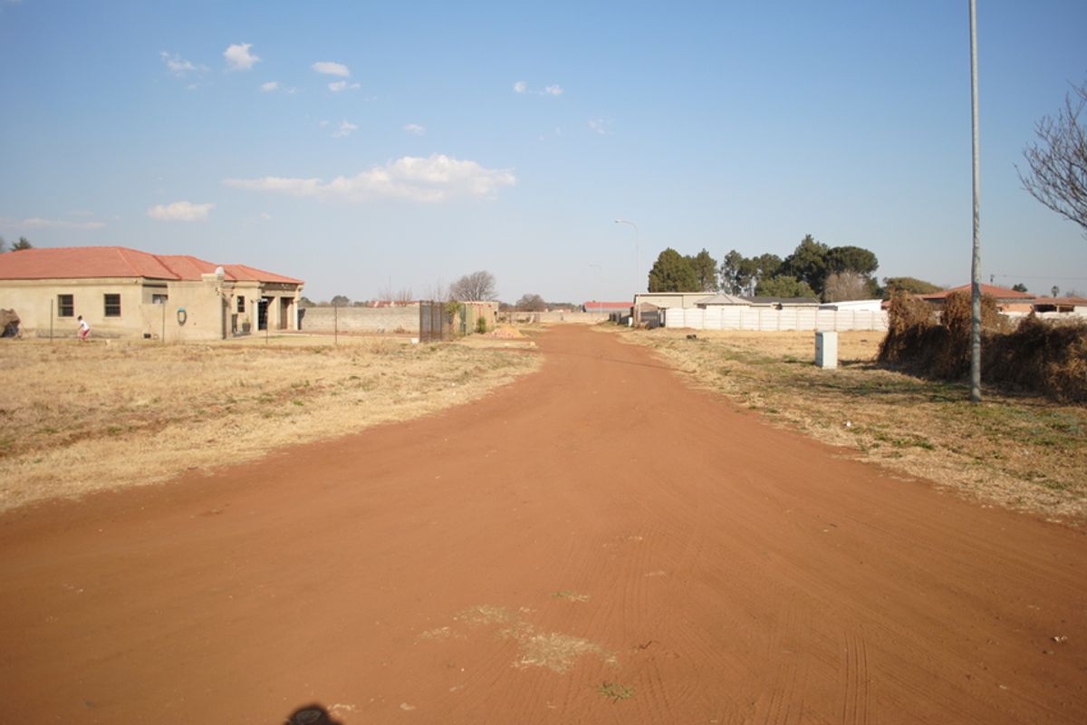 A view of an unpaved residential street showing surrounding properties and perimeter fencing in a developing neighborhood.