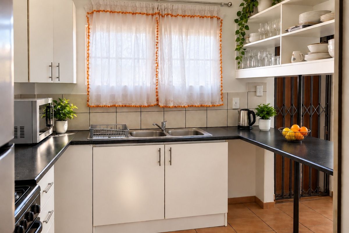 A functional kitchen featuring white cabinetry, dark laminate countertops, a double bowl stainless steel sink, and open shelving with tiled flooring.
