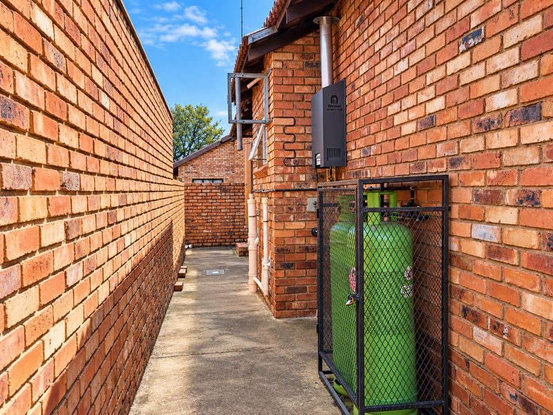 Side exterior area featuring a brick facade, concrete pathway, wall-mounted gas water heater, and a secure metal cage for gas cylinders.