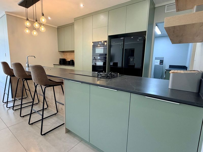 Modern open-plan kitchen featuring sage green cabinetry, black stone countertops, built-in appliances, and a breakfast bar with pendant lighting.