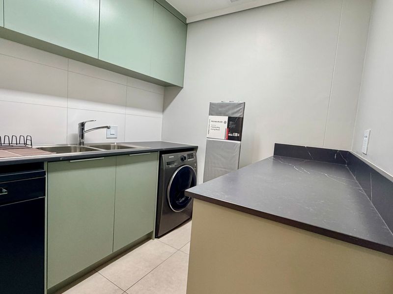 A modern, well-maintained laundry room featuring sage green cabinetry, dark stone-look countertops, a stainless steel sink, and tiled flooring.