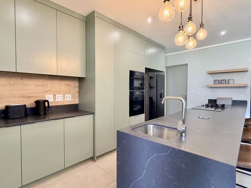 Modern kitchen featuring sage green cabinetry, a large stone island with an undermount sink, built-in appliances, and contemporary pendant lighting.