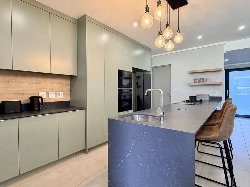 Modern kitchen featuring sage green cabinetry, a dark stone island with integrated sink, built-in appliances, and contemporary pendant lighting.
