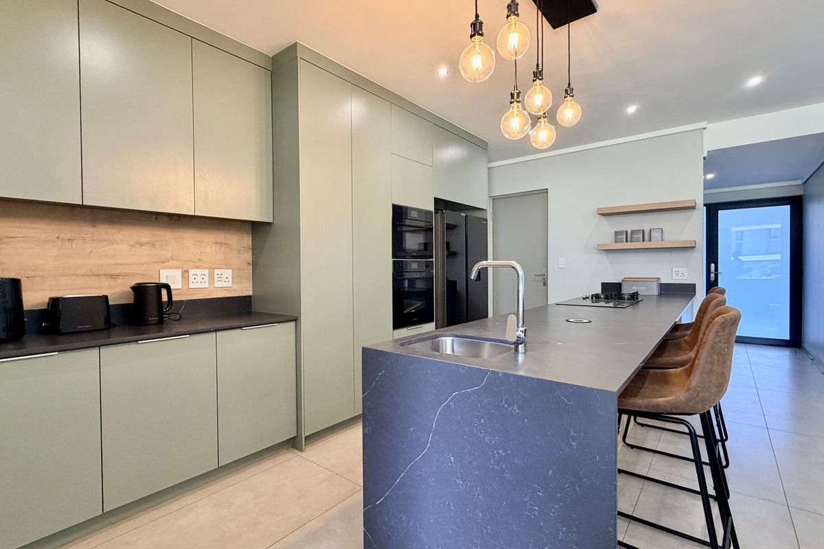 Modern kitchen featuring sage green cabinetry, a dark stone island with integrated sink, built-in appliances, and contemporary pendant lighting.