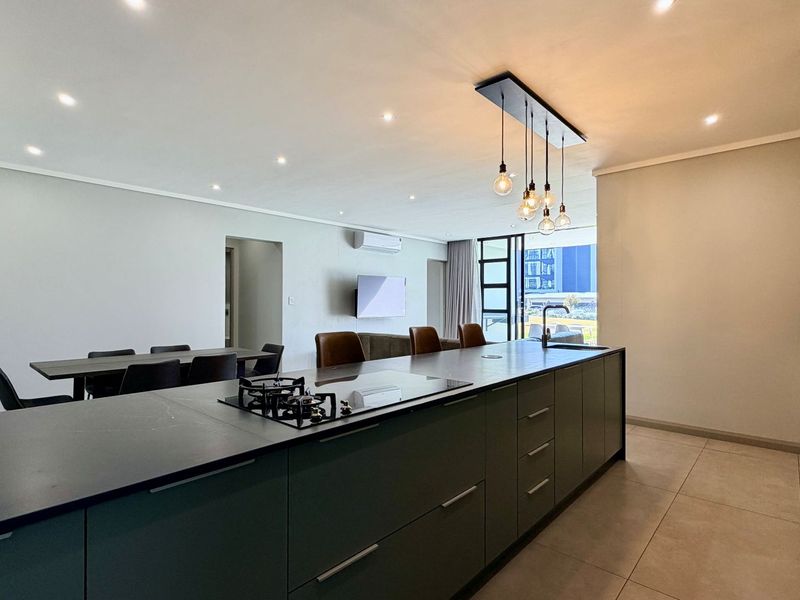 Modern open-plan living area featuring a sleek kitchen island with gas hob, integrated sink, and contemporary pendant lighting, flowing into the dining and lounge space.
