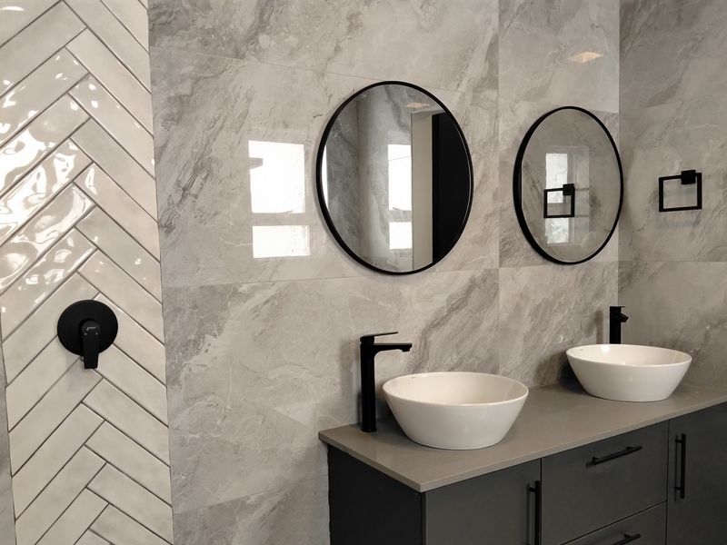 Modern bathroom featuring a double vanity with vessel sinks, matte black fixtures, and elegant grey marble-look wall tiles with a herringbone accent.