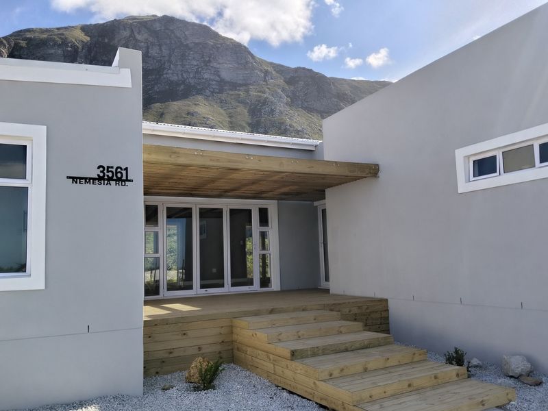 Modern front entrance featuring timber decking, steps, and a pergola, complemented by clean grey exterior walls and mountain views.