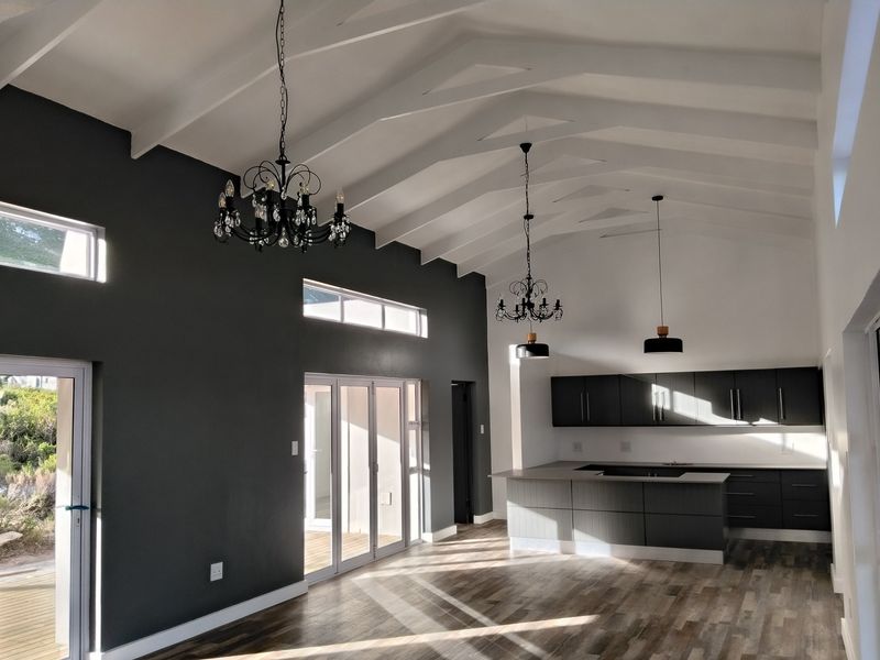 Spacious open-plan living and kitchen area featuring vaulted ceilings with exposed rafters, modern dark cabinetry, a central island, and large sliding doors to the exterior.