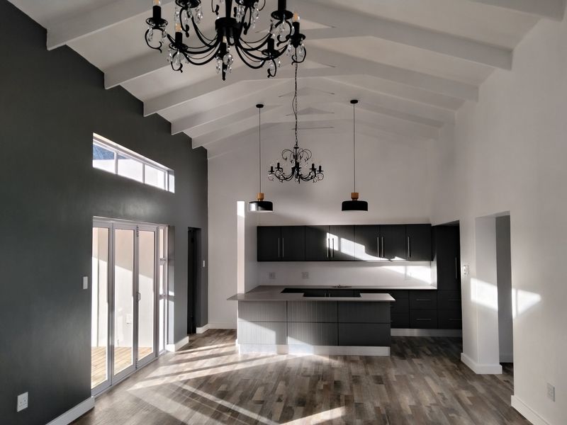 Spacious open-plan living and kitchen area featuring a high vaulted ceiling with exposed beams, modern dark cabinetry, and large bi-fold doors leading outdoors.