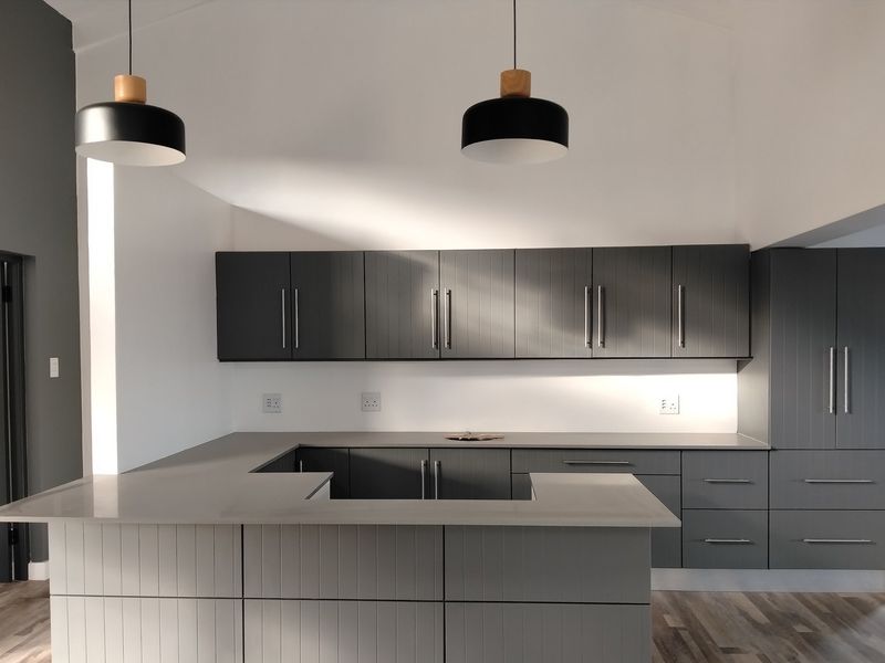 A modern, minimalist kitchen featuring sleek grey cabinetry, stone countertops, and contemporary pendant lighting in a bright, open-plan space.