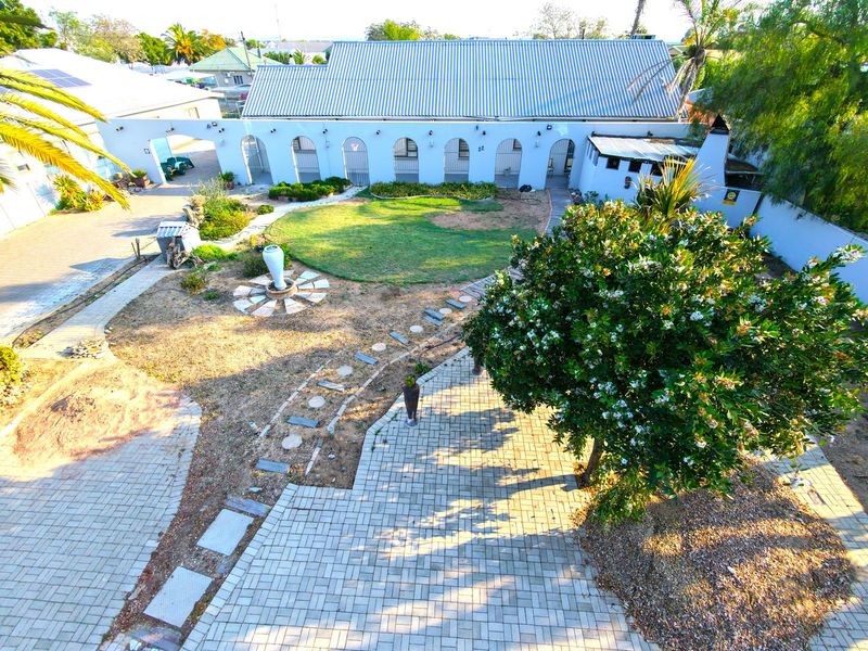 Aerial view of a property featuring a large corrugated metal roof, arched architectural details, a paved driveway, and a landscaped garden area.