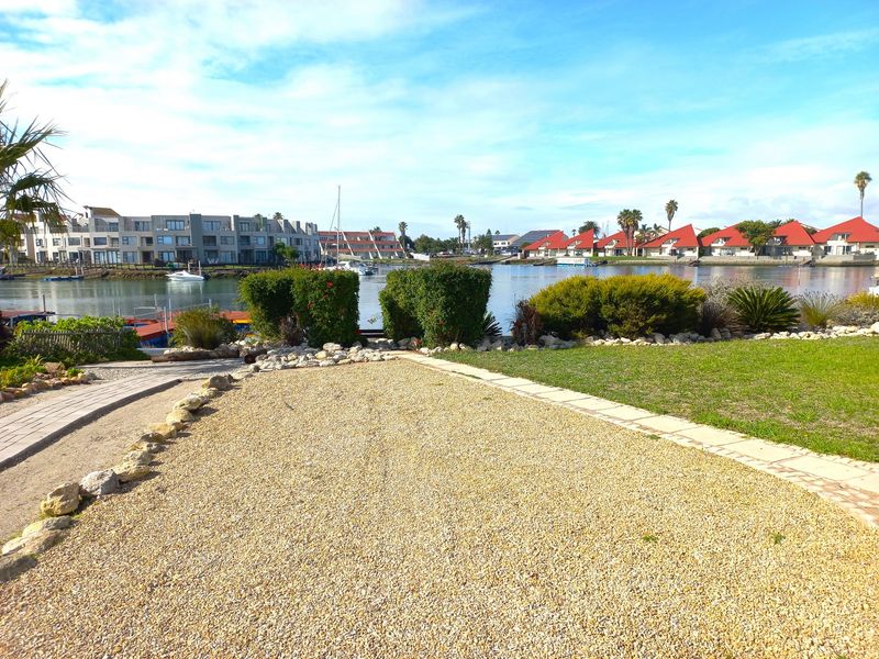 A scenic view of the waterfront residential area featuring a well-maintained garden, gravel driveway, and paved pathways overlooking the canal.