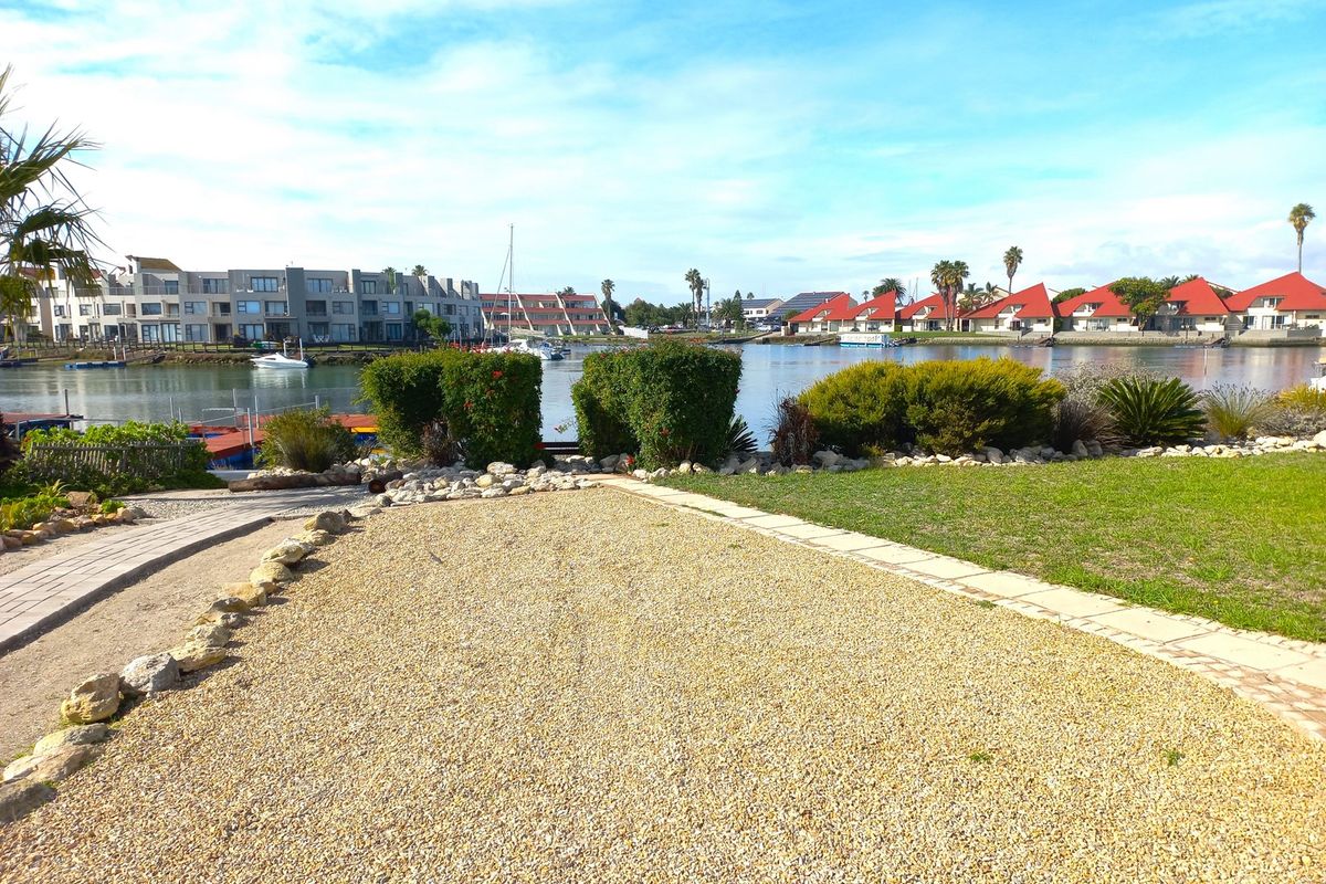 A scenic view of the waterfront residential area featuring a well-maintained garden, gravel driveway, and paved pathways overlooking the canal.