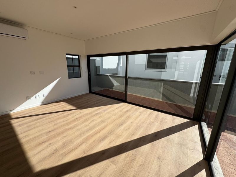 A bright, modern, empty bedroom featuring light wood-look laminate flooring, recessed lighting, a split-system air conditioner, and large sliding glass doors opening to a balcony.