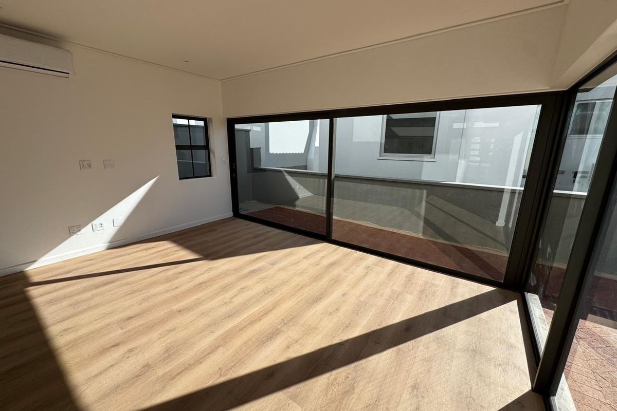 A bright, modern, empty bedroom featuring light wood-look laminate flooring, recessed lighting, a split-system air conditioner, and large sliding glass doors opening to a balcony.