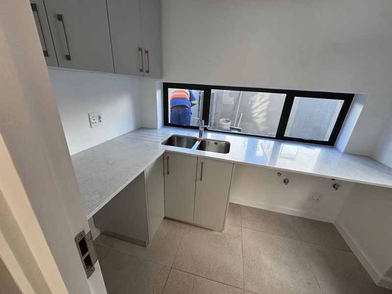 A modern, clean scullery featuring stone countertops, a double basin sink, and ample built-in cabinetry with neutral tiled flooring.