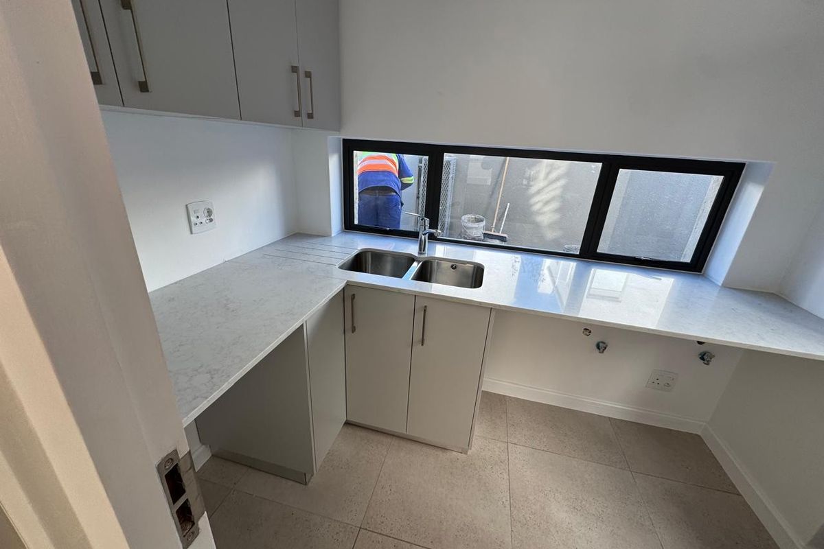 A modern, clean scullery featuring stone countertops, a double basin sink, and ample built-in cabinetry with neutral tiled flooring.