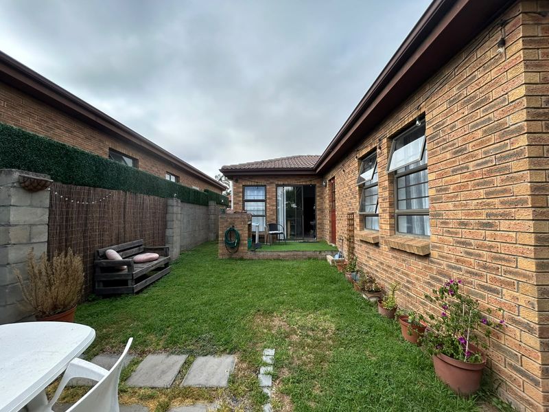 A private rear garden featuring a face brick townhouse exterior, a small paved patio, and a lawn area enclosed by boundary walls.