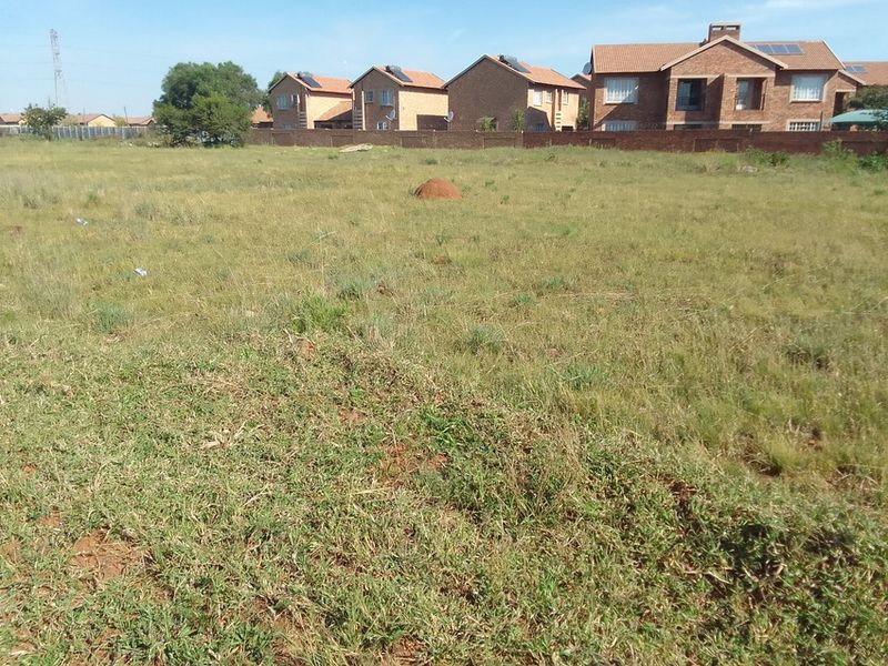 A vacant residential plot of land featuring natural grass cover and a perimeter wall, situated in a developed residential neighborhood.