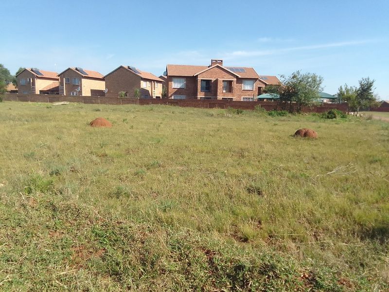 A vacant residential plot of land featuring natural grassland, enclosed by a perimeter wall with neighboring residential properties visible in the background.