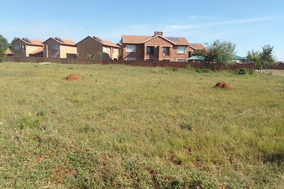 A vacant residential plot of land featuring natural grassland, enclosed by a perimeter wall with neighboring residential properties visible in the background.