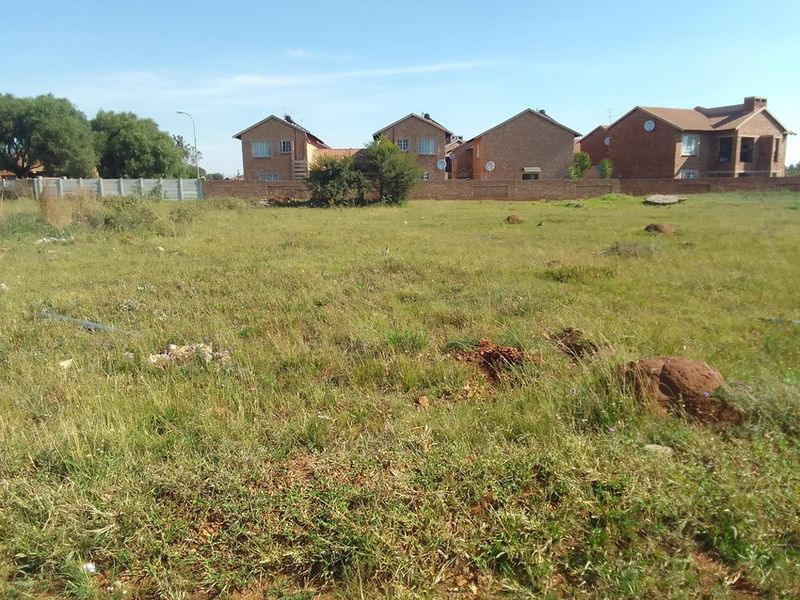 A vacant residential plot of land featuring natural grass cover and a perimeter boundary wall, situated in a developed residential neighborhood.