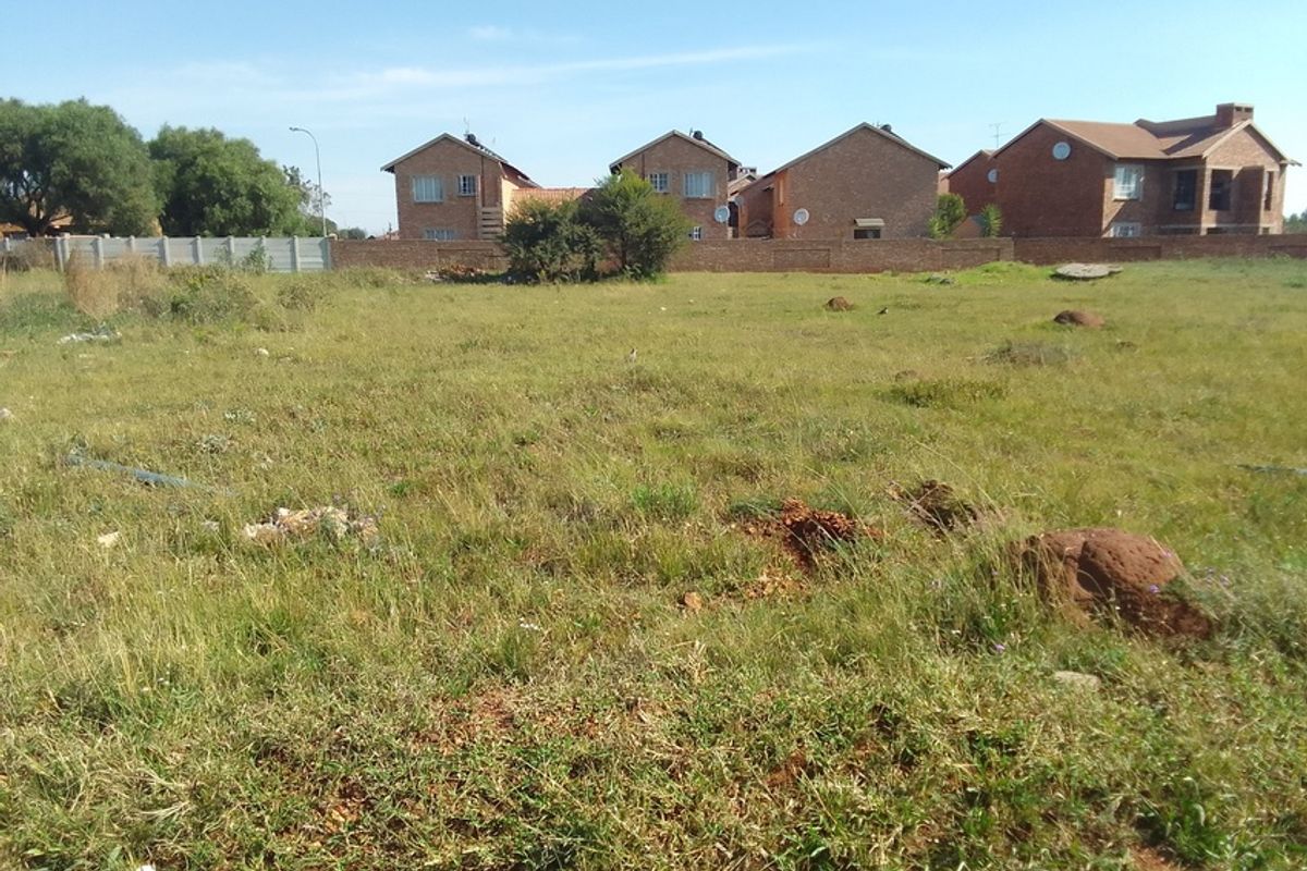 A vacant residential plot of land featuring natural grass cover and a perimeter boundary wall, situated in a developed residential neighborhood.