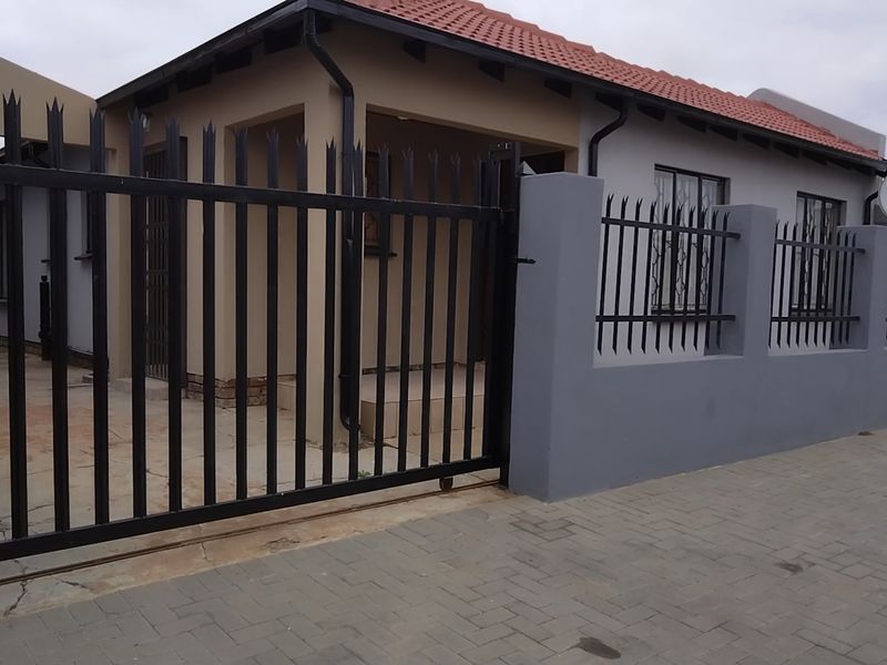 Front exterior view of a residential property featuring a secure metal gate, perimeter wall with security spikes, and a paved driveway.