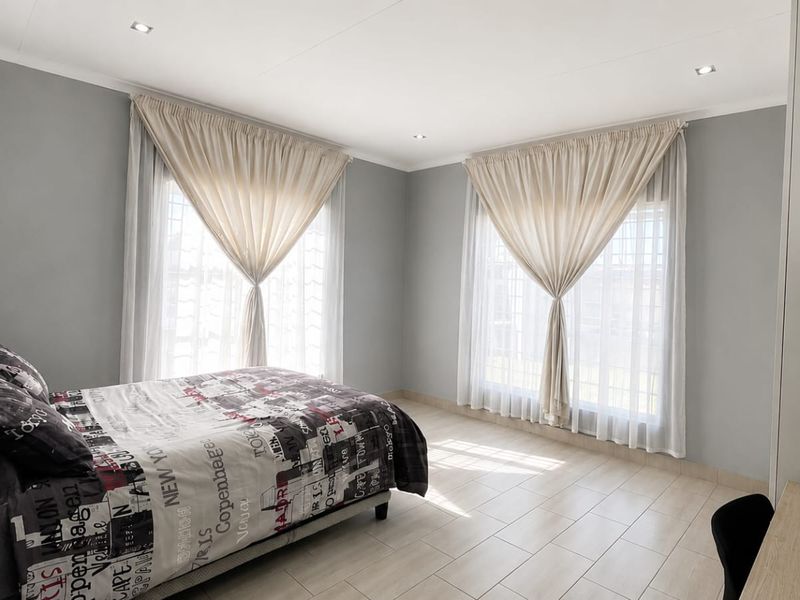 A bright and spacious bedroom featuring neutral grey walls, tiled flooring, and large windows with elegant drapery, creating a clean and comfortable living space.
